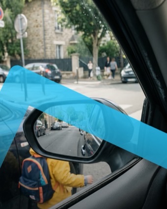 Vue depuis le poste de conduite : dans le rétroviseur, un enfant en manteau jaune passe derrière la voiture
